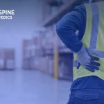 Worker in a safety vest gripping his lower back in a warehouse